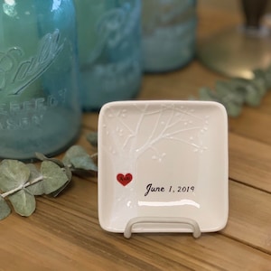 May include: White ceramic ring dish with a tree design and a red heart with the initials "A & M" and the date "June 1, 2019".