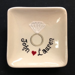 May include: White ceramic ring dish with a raised diamond ring design and the names "John" and "Lauren" written in black cursive with a red heart between the names.