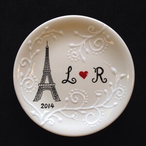 May include: White ceramic plate with a black and white illustration of the Eiffel Tower, a red heart, and the letters "L" and "R". The plate also has a white floral design and the year "2014" printed on it.