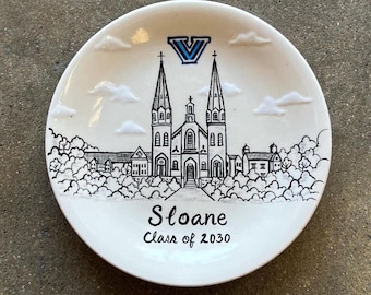 May include: A white ceramic plate featuring a black and white illustration of a church with two spires, clouds, and trees. The plate has the text "Sloane Class of 2030" and a blue and white logo at the top.