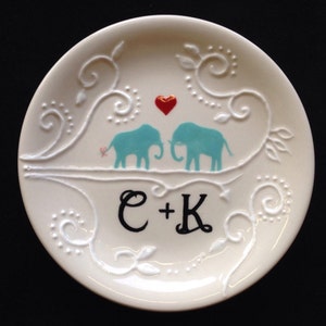 May include: White ceramic ring dish with a raised design of two elephants facing each other with a red heart above them. The dish has a white floral design and the initials "C + K" in black.