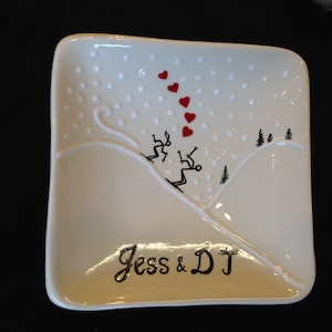 Puede incluir: Plato de cerámica blanco con una escena de montaña nevada y esquiadores. El plato está decorado con corazones rojos y el texto "Jess & DJ" en negro.