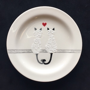 May include: White ceramic plate with two black and white cats facing each other with a red heart above them. The cats have a swirl pattern on their bodies and are sitting on a gray line.