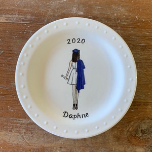 May include: White ceramic plate with a blue and white illustration of a graduate in a cap and gown. The plate has a white rim with small white dots. The text "2020" and "Daphne" are written on the plate.