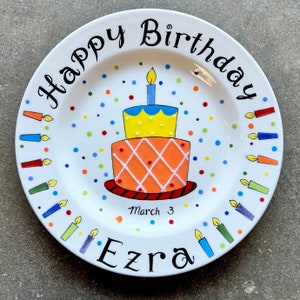 May include: A white ceramic plate with a hand-painted birthday cake design. The cake is orange and yellow with a blue candle on top. The plate is decorated with colorful confetti and candles around the edge. The text "Happy Birthday Ezra March 3" is written on the plate.