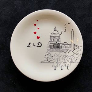 May include: A white ceramic plate featuring a black and white illustration of the US Capitol building and the Washington Monument. The plate has red hearts in the top left corner and the initials "L & D" in the centre.
