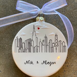 May include: White ceramic Christmas ornament with a black and white illustration of the Chicago skyline. The ornament has a red heart above the skyline and the names "Nik & Megan" written below.