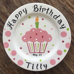 May include: A white ceramic plate with a pink cupcake design, a lit candle, and the words "Happy Birthday" and "Tilly". Pink and green polka dots surround the plate. The date "May 23" is also included.
