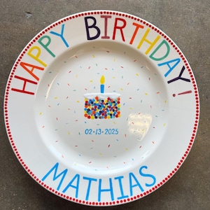 May include: White ceramic plate with a colorful "Happy Birthday!" message and the name "Mathias" in blue. The plate features a cake illustration with a lit candle and confetti. The date "02-13-2025" is also printed on the plate.