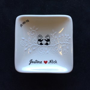May include: White ceramic ring dish with a raised design of two pandas holding hands on a branch. The dish is inscribed with the date "12/10/16" and the names "Justina" and "Nick" with a red heart symbol between them.