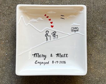 May include: A square white ceramic trinket dish featuring a raised mountain outline, two stick figures hiking, five red hearts, two black birds, and a sign reading Lake Placid. The text reads Mary & Matt Engaged 8-17-2026 in black script.