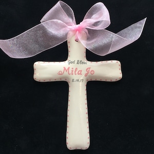 May include: A white ceramic cross with pink polka dots and the inscription "God Bless Mila Jo 5.14.17." A sheer pink ribbon is tied at the top. The cross is a religious item, suitable for a baptism or christening.