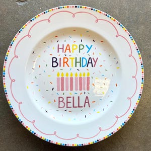 May include: White ceramic plate with a pink scalloped border and colorful polka dots. The plate features the words "HAPPY BIRTHDAY BELLA" in various colors, with candles and confetti.