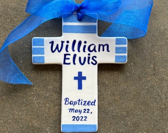 May include: A white ceramic cross with blue stripes and the name "William Elvis" written on it. The cross also has a small cross symbol and the text "Baptized May 22, 2022".