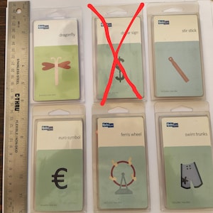 Puede incluir: Seis paquetes de plástico transparente con etiquetas verdes y blancas que contienen formas troqueladas para manualidades. Las formas incluyen una libélula, un signo de dólar, un palo para remover, un símbolo de euro, una rueda de la fortuna y un bañador.