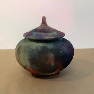 May include: A handmade ceramic jar with a lid. The jar has a round, bulbous body and three small feet. The lid features a knob. The jar is painted with a mix of colours, including blue, green, purple, and red.