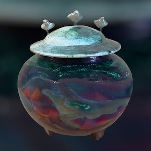 Puede incluir: Un jarrón de cerámica con tapa, que presenta una mezcla de colores que incluyen azul, verde y rojo. El jarrón tiene un cuerpo redondo con tres patas pequeñas y está coronado por una tapa adornada con tres elementos decorativos plateados.