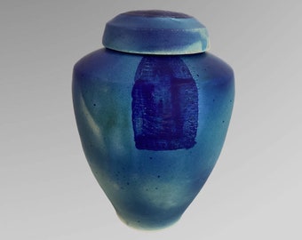Op de afbeelding: Een blauwe keramische pot met een deksel. De pot heeft een afgeronde body en een donkerblauw rechthoekig ontwerp aan de voorkant. Het deksel is een lichtere tint blauw met een textuur.