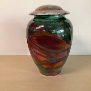 May include: A large, handcrafted ceramic urn with a lid. The urn has a glossy finish with a blend of colours, including deep green, brown, and hints of iridescent hues. The design includes flowing patterns and a rounded shape, suitable for home decor.