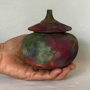May include: A small, round ceramic jar with a lid. The jar is decorated with a colourful, abstract design in shades of green, red, and purple. The jar has three small feet.
