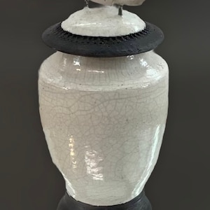 May include: A white ceramic jar with a black base and lid. The lid is topped with a decorative element featuring three small white bird figurines. The jar has a crackle glaze finish, adding texture to the piece.