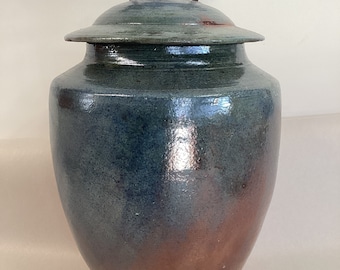 May include: A handcrafted ceramic urn with a lid, featuring a gradient of blue and copper hues. The urn is adorned with a decorative element of three small bird figures perched on a branch. The piece has a glossy finish and a unique, artistic design.
