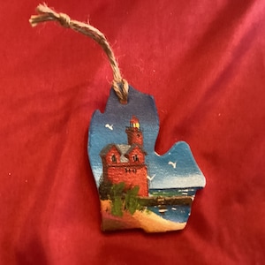 May include: A hand-painted wooden ornament shaped like the state of Michigan with a red lighthouse on a blue lake. The ornament has a twine hanger.