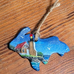 May include: Hand-painted ornament in the shape of a fish, featuring a lighthouse scene. The ornament has a blue background with a lighthouse, a red-roofed building, and a jute string for hanging. The lighthouse has a red and yellow top.