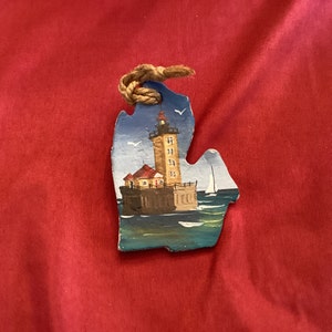 May include: A hand-painted wooden ornament shaped like the state of Michigan, featuring a lighthouse with a red roof and a blue sky background.