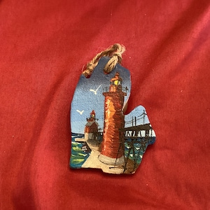 May include: A hand-painted wooden ornament shaped like the state of Michigan, featuring a red lighthouse with a white light on a pier. The ornament is painted with blue water, a blue sky, and white birds.