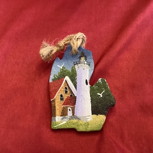 May include: A hand-painted wooden ornament shaped like the state of Michigan, featuring a white lighthouse with a red roof and a yellow house with a red roof. The ornament has a brown twine hanger.