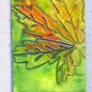 May include: A rectangular artwork featuring a stylized leaf design. The leaf is rendered in shades of orange, yellow, and green, with a textured appearance. The background is a watercolor wash of green.