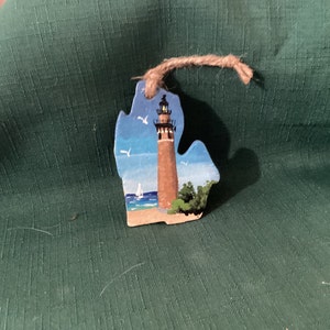 May include: A wooden ornament shaped like the state of Michigan with a lighthouse painted on it. The lighthouse is brown and white with a blue roof and a light on top. The background is a blue sky with white clouds and a blue lake with a sailboat.