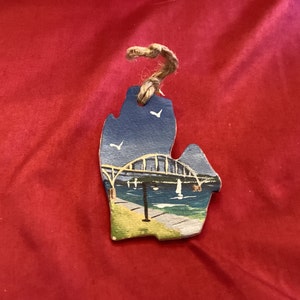 Może przedstawiać: Drewniana ozdoba w kształcie stanu Michigan, ręcznie malowana z widokiem na most pod błękitnym niebem. Ozdoba ma pętlę z juty do zawieszenia. Tło to czerwona tkanina.