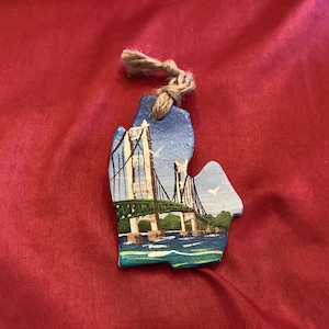 May include: A hand-painted wooden ornament shaped like the state of Michigan with a depiction of the Mackinac Bridge in blue, green, and white.