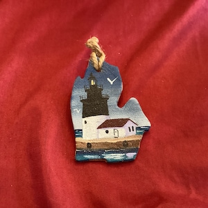 Könnte beinhalten: Ein handbemalter Holzschmuck in Form des Bundesstaates Michigan mit einem Leuchtturm auf einem blauen See. Der Leuchtturm ist weiß mit einem schwarzen Turm und einem roten Dach. Ein kleiner Vogel fliegt über dem Leuchtturm.