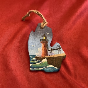 May include: Wooden ornament in the shape of the state of Michigan, featuring a lighthouse scene. The lighthouse is painted red with a black top, set against a blue sky and ocean. A jute string is attached for hanging.