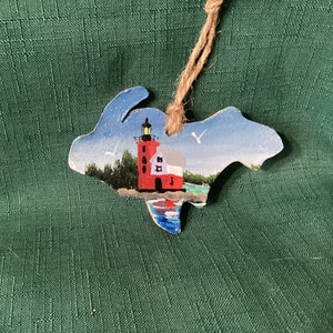 May include: A hand-painted wooden ornament shaped like the state of Michigan with a red lighthouse and a white house on a blue lake.