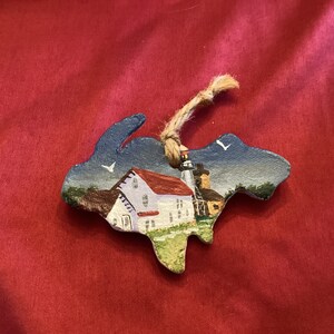 Puede incluir: Un adorno de madera pintado a mano con forma del estado de Michigan, que presenta una casa blanca con techo rojo y un faro en el fondo. El adorno tiene un colgador de cuerda rústico.