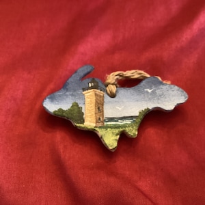 May include: A wooden ornament shaped like the state of Michigan with a painted scene of a lighthouse on a lake shore. The ornament has a brown twine hanger.
