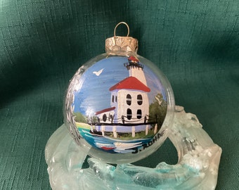 Op de afbeelding: Een handgeschilderde glazen ornament met een vuurtoren met een rood dak en witte muren. De vuurtoren is geschilderd op een blauwe achtergrond met een witte vogel die erboven vliegt. De ornament is onderaan gesigneerd met "J. Roha".