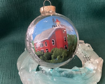 May include: A glass Christmas bauble featuring a painted image of a red lighthouse with white trim and a white roof. The lighthouse is surrounded by green trees and a blue sky with white clouds. The bauble is sitting on a clear glass base.