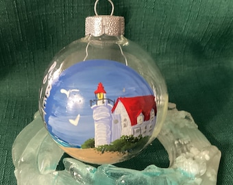 May include: Hand-painted glass bauble depicting a white lighthouse with a red roof set against a blue sky. The lighthouse is situated on a sandy beach with a small green bush in front. Two white birds are flying in the sky. The bauble is made of clear glass with a silver top.