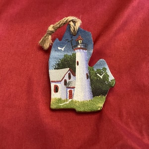 May include: A hand-painted wooden ornament shaped like the state of Michigan, featuring a white lighthouse with a red roof and a small white house with a red door. The ornament is painted on a blue background with green trees and white birds.