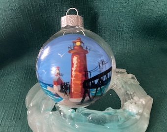 May include: A hand-painted glass bauble depicting a red lighthouse with a white light on top, a pier, and a blue sky with a white bird. The bauble is resting on a clear glass stand.