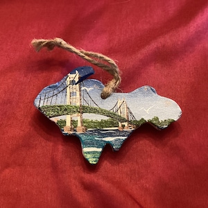 Może przedstawiać: Drewniana ozdoba ręcznie malowana w kształcie stanu Michigan z niebieskim mostem namalowanym na górze. Ozdoba ma brązowy sznurek do zawieszenia.