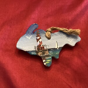 May include: A hand-painted wooden ornament shaped like a fish, featuring a lighthouse scene. The lighthouse is red and white, set against a blue sky with seagulls. A natural twine loop is attached for hanging.