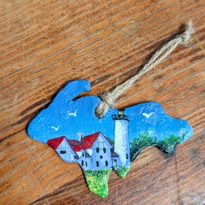 May include: Hand-painted wooden ornament shaped like a state, featuring a lighthouse and buildings with red roofs. The ornament has a blue sky background with white birds and a jute string for hanging.