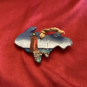 May include: A hand-painted wooden ornament shaped like the state of Michigan with a red lighthouse and a sailboat on a blue lake. The ornament has a brown twine string for hanging.