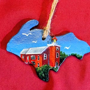May include: Hand-painted wooden ornament in the shape of a state, featuring a red lighthouse with a white roof and a blue sky with white birds. The ornament is suspended by a natural twine.